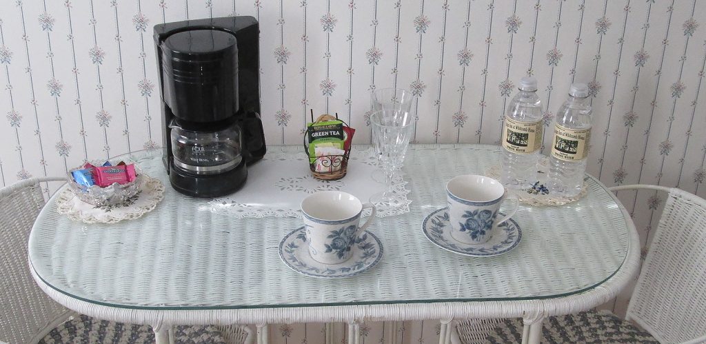 Wicker table and chairs with coffee maker, supplies, 2 tea cups, bottled water, 2 wine glasses and bowl of chocolates