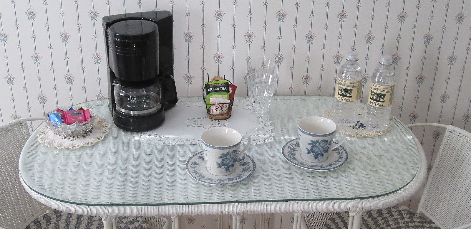 Wicker table with coffee maker, tea cups, bottled water, and a bowl of chocolates